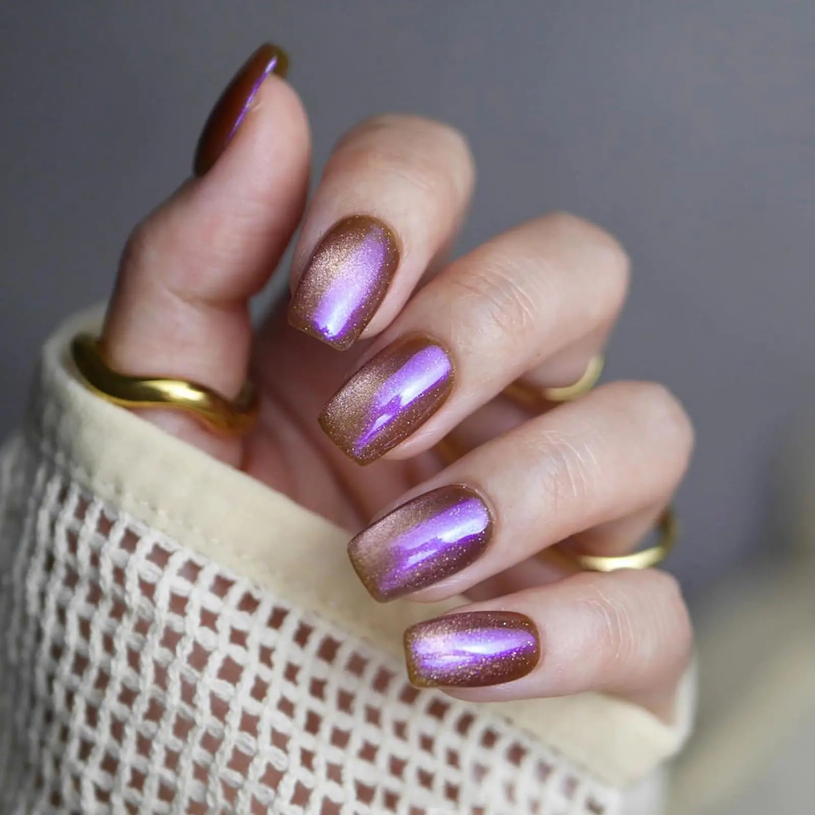 Hand with purple and gold nail polish wearing a gold ring on a neutral background
